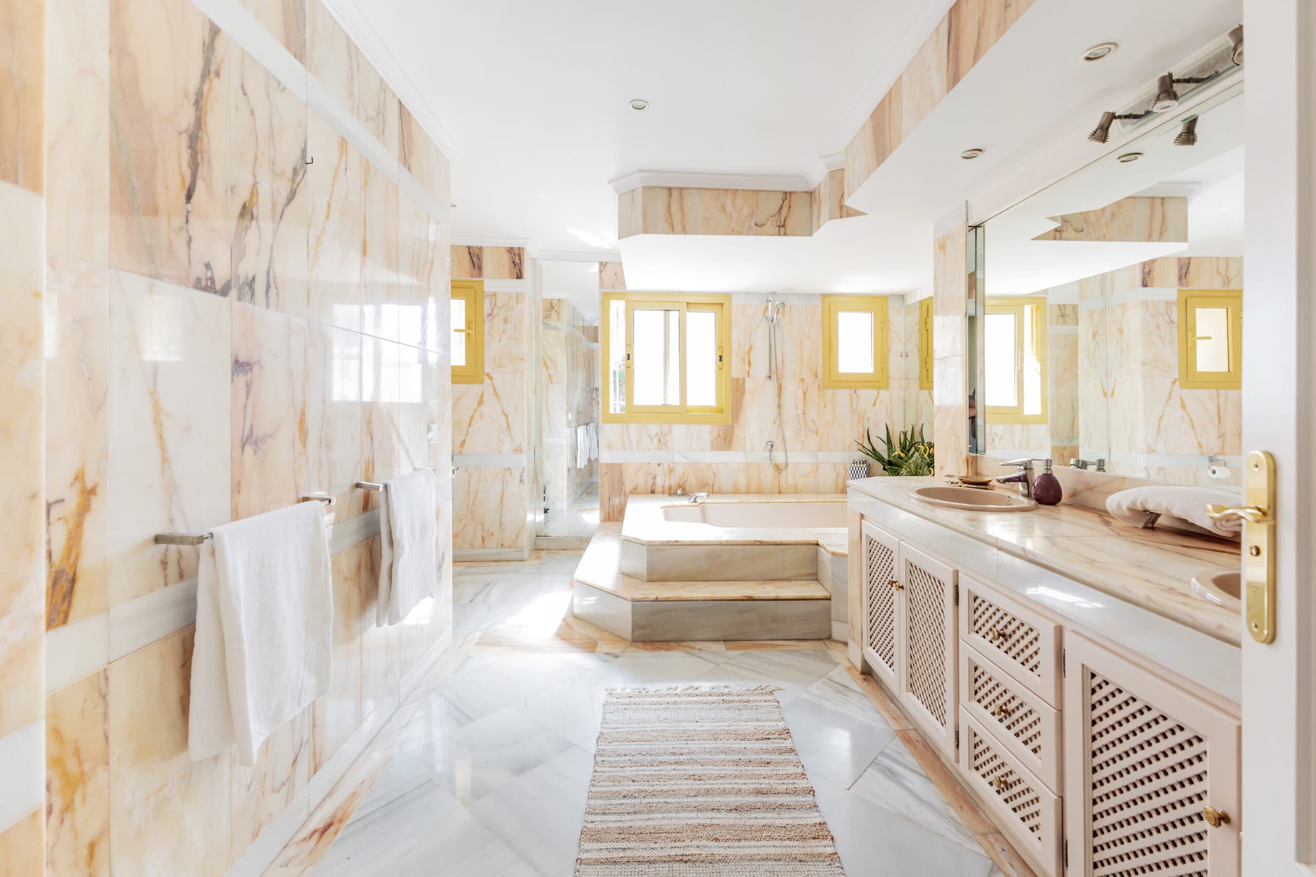 A long marble bathroom with a raised bath and sunlit yellow-framed windows